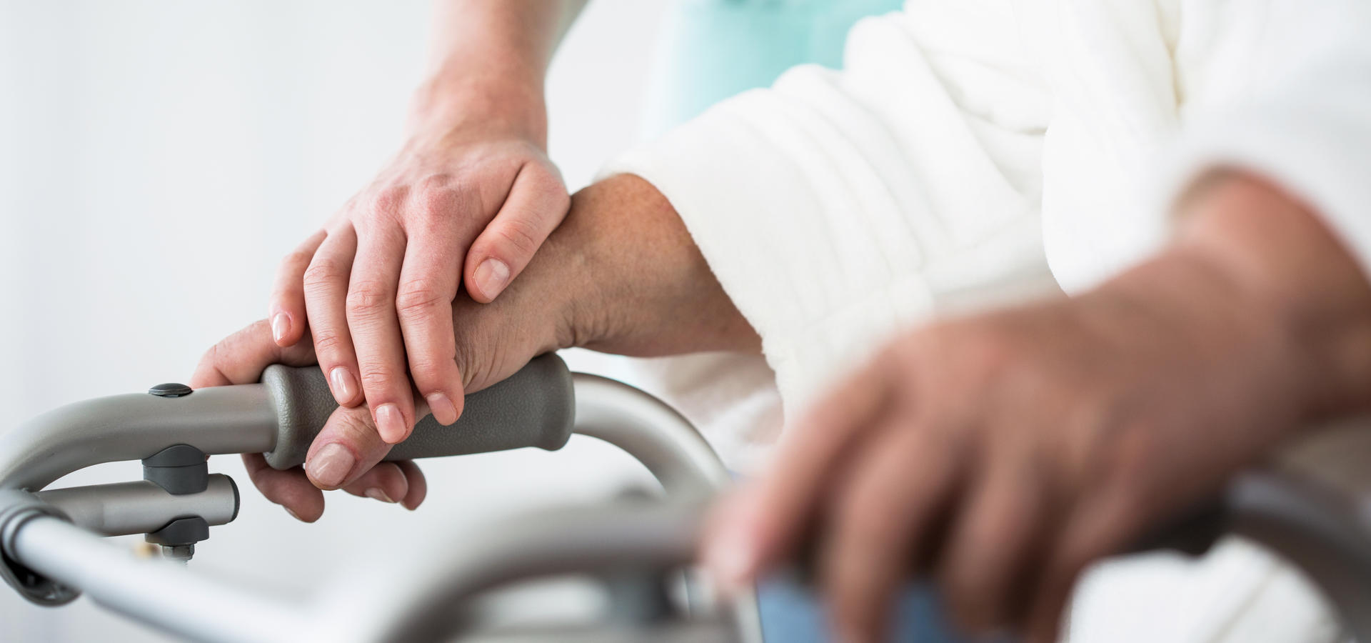 A senior living and long-term care themed photo: a caregiver's hand gently rests on one of the hands of a senior living resident, whose hands are holding on to a walker. A close up photo. 
