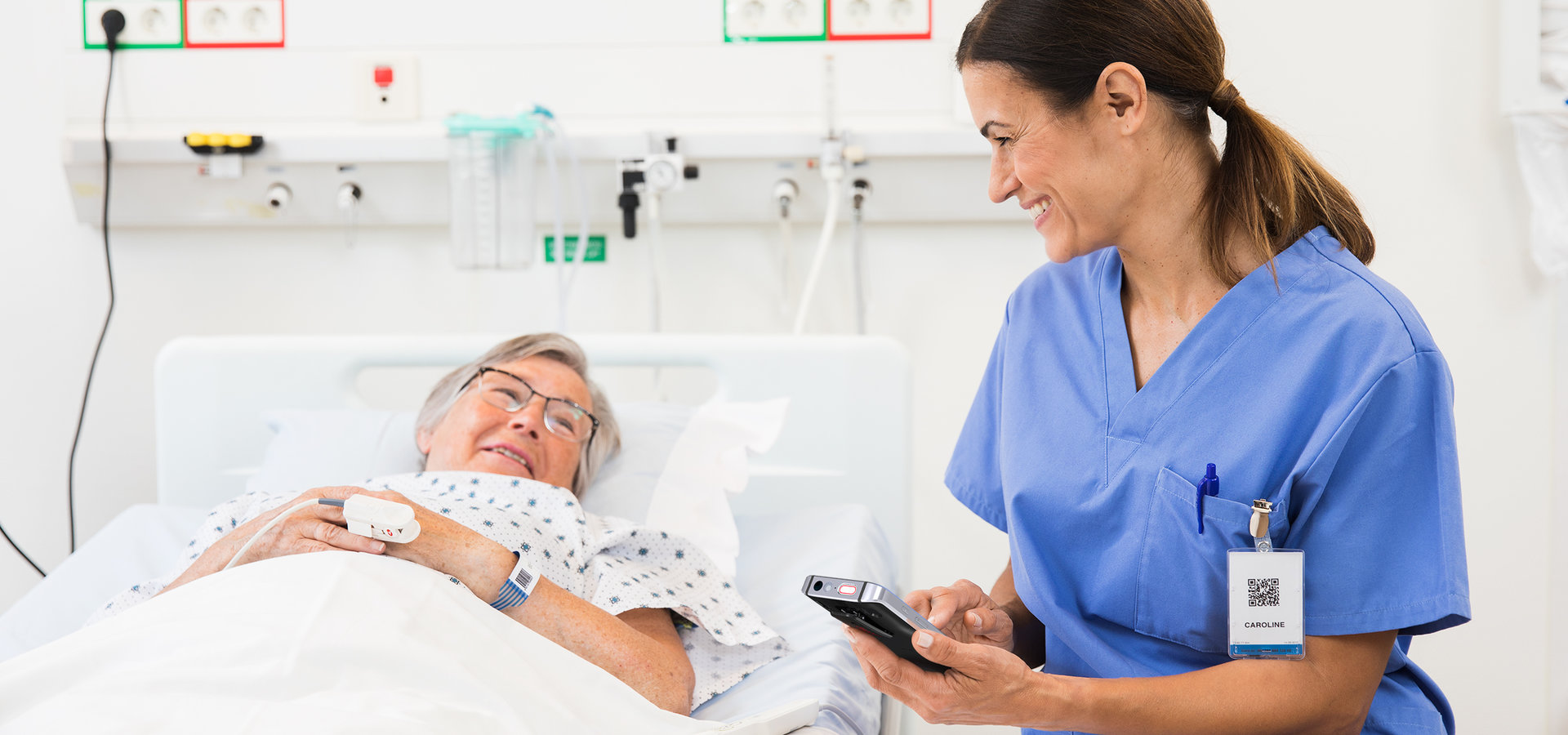 A medical doctor or a nurse with a patient, by the patient's hospital bed. Both the patient and the nurse are smiling and probably in a conversation; the nurse is holding and typing on a nurse call or communication system device.