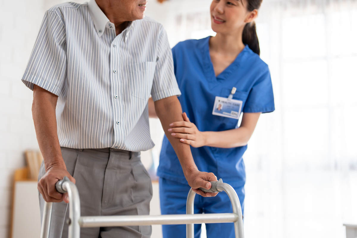 Senior living themed photo: male resident with a walker being assisted by a nurse or a care worker in blue at a senior home or long term health facility.